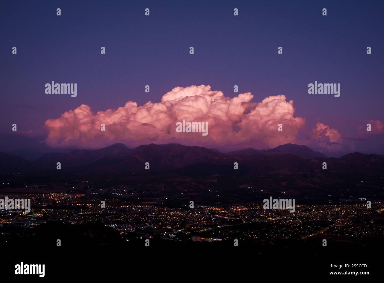 Hochperspektivisches Landschaftsfoto des frühen Nachthimmels mit einer flauschigen violetten Wolke, die über einer Stadt mit Lichtern hängt, gibt es auch eine Bergkette. Stockfoto