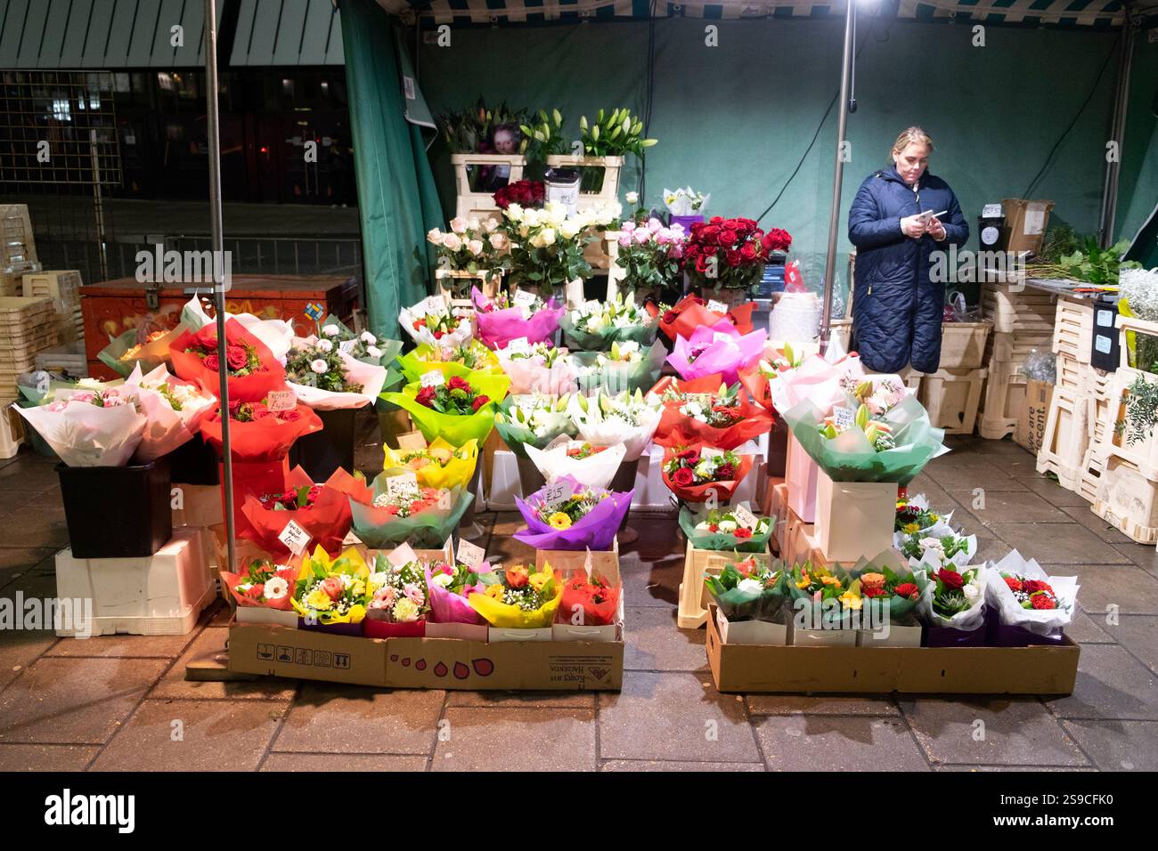 Ältere Frau, die im Winter an einem Januarabend in Zentral-London England Großbritannien 2025 Großbritannien KATHY DEWITT auf der Straße Schnittblumen verkauft Stockfoto