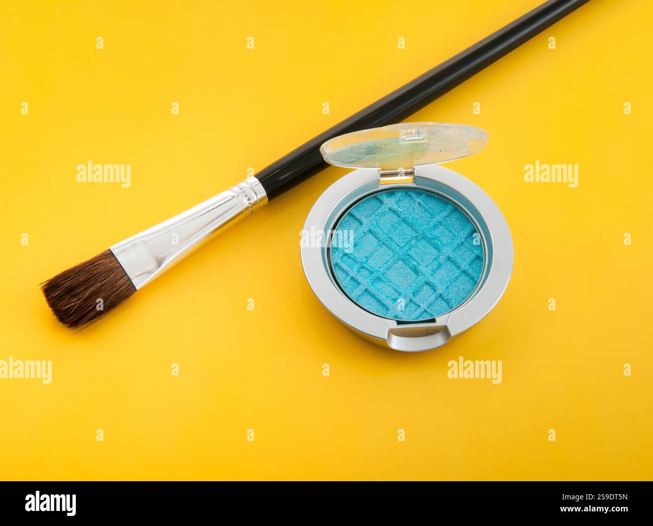 Ein auffälliger flacher Lay mit einem blau gepressten Lidschatten und einem Make-up-Pinsel, alles auf einem kräftigen, leuchtend gelben Hintergrund. Pro Stockfoto