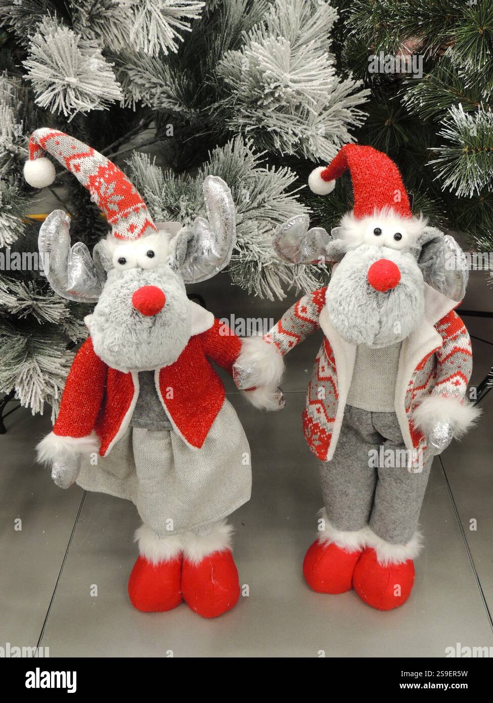 Ein Paar lustige Elche oder Hirsche in Weihnachtspullover und Hüten stehen unter einem silbernen künstlichen Weihnachtsbaum und halten sich an den Pfoten Stockfoto