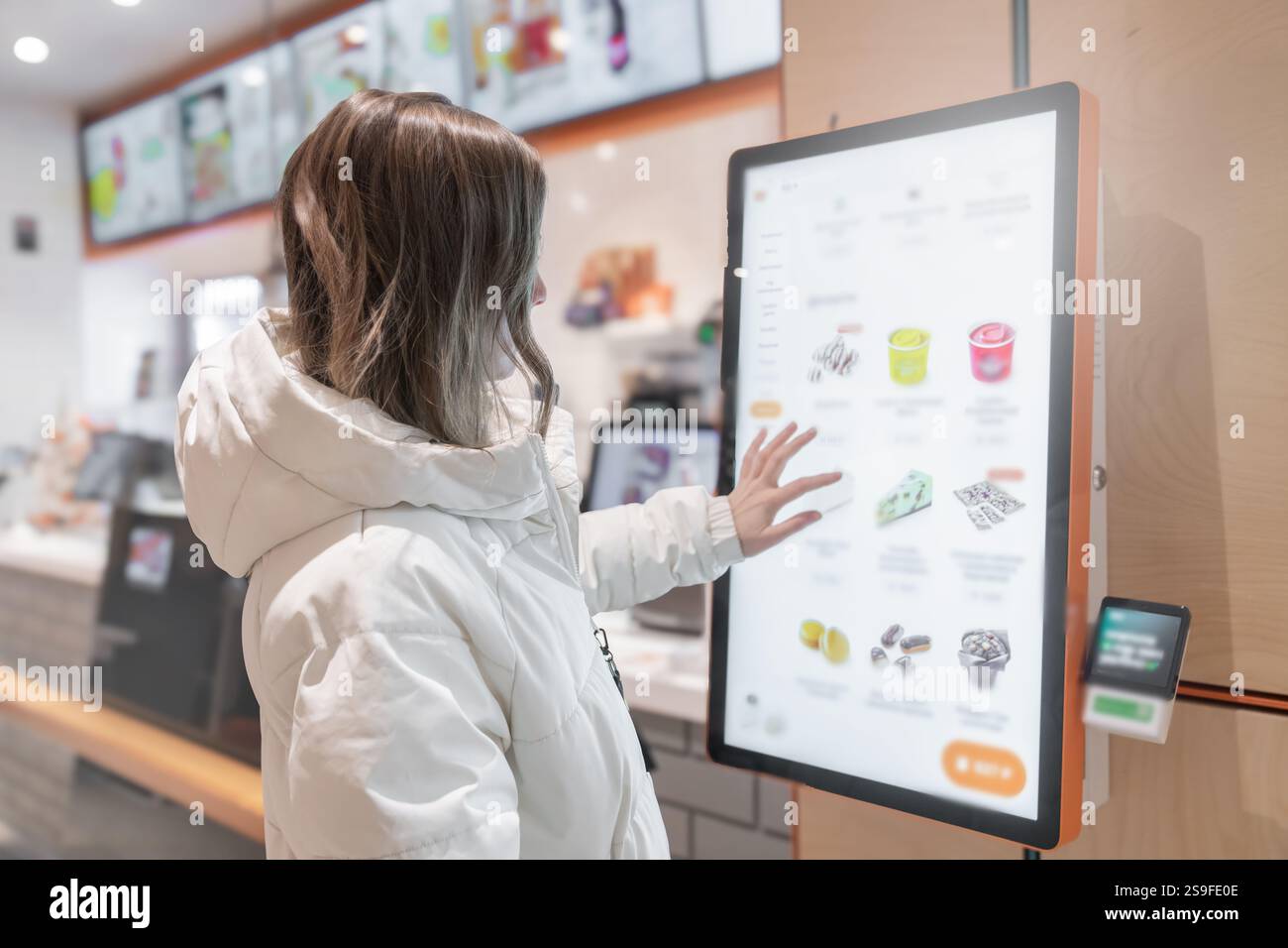 Eine 30-35-jährige Frau in einer weißen Winterjacke bestellt Essen über die Bildschirmkarte in einem Fast-Food-Restaurant. Eine junge Person nutzt aktiv Stockfoto