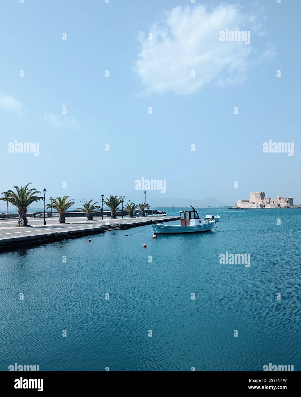 Ruhiger Blick auf das Schloss Bourtzi in Nafplio, mit einem einsamen Boot, das auf ruhigem blauem Wasser schwimmt, und einer palmengesäumten Promenade im Vordergrund. Stockfoto