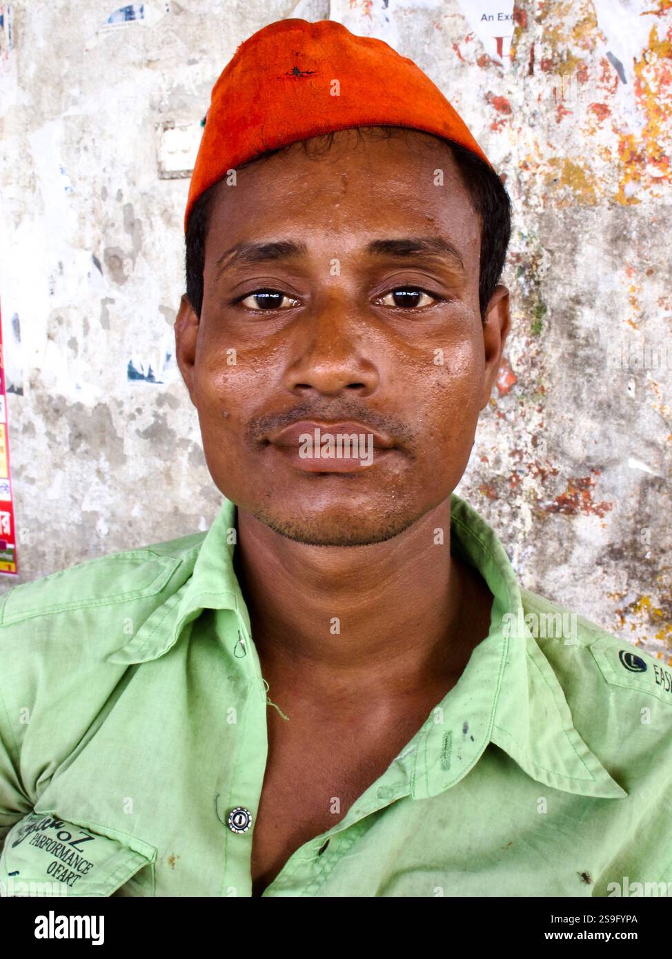 Porträt eines Mannes mit orangefarbener Mütze und grünem Hemd, aufgenommen in Bangladesch, mit einem ruhigen Ausdruck vor einer strukturierten Wand mit verblassten Plakaten. Stockfoto