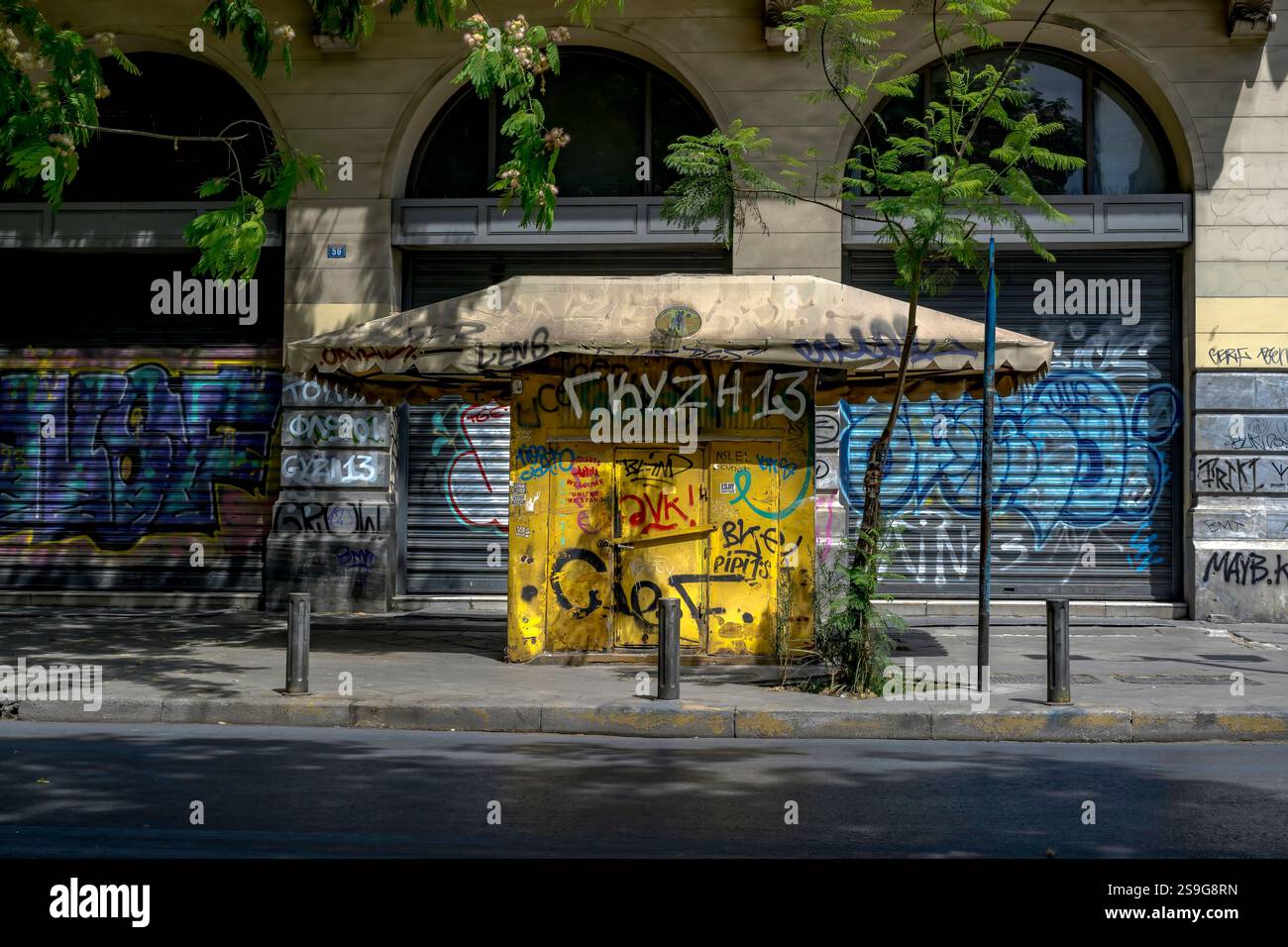 Ein kleiner, gelber Stand mit farbenfrohen Graffiti, in einer städtischen Straße gelegen, umgeben von verschlossenen Geschäften, die auch mit Graffiti geschmückt sind, die sich spiegeln Stockfoto