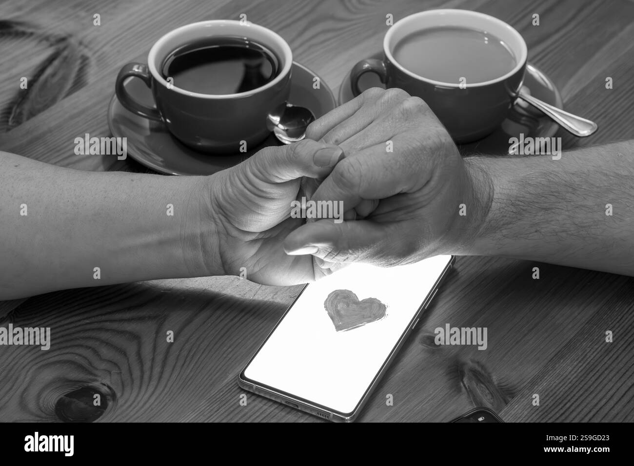 Das Bild zeigt ein Paar, das mit zwei Tassen Kaffee an einem Tisch sitzt. Ihre Hände sind zusammengeschnallt, und zwischen ihnen befindet sich ein Smartphone mit einem R Stockfoto