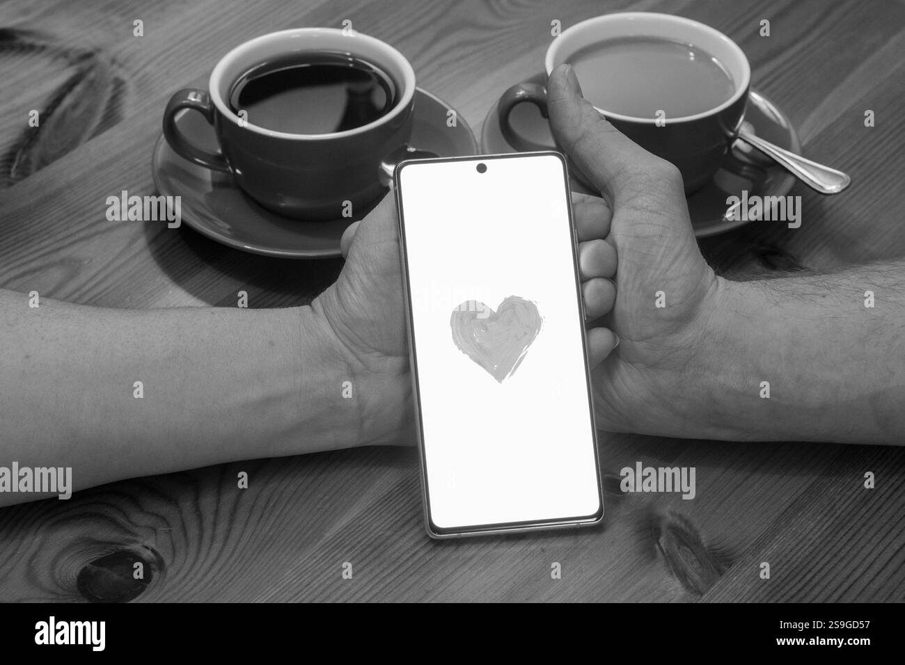 Das Bild zeigt ein Paar, das mit zwei Tassen Kaffee an einem Tisch sitzt. Ihre Hände sind zusammengeschnallt, und zwischen ihnen befindet sich ein Smartphone mit einem R Stockfoto