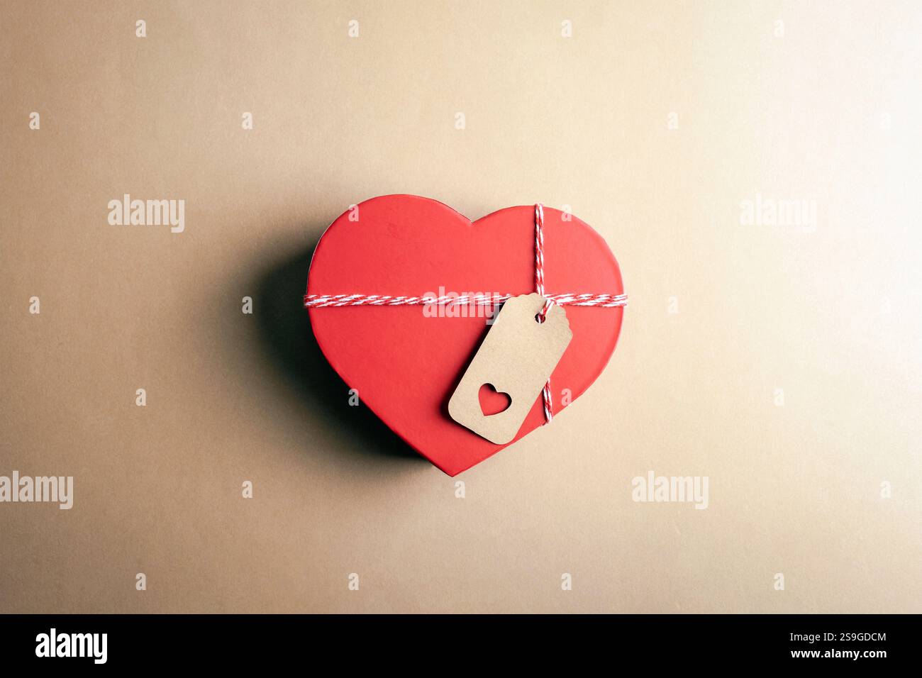 Rote Box in Herzform mit einem Kraftanhänger auf beigefarbenem Hintergrund, das Liebe und nachdenkliches Geschenk darstellt. Valentinstag-Konzept. Draufsicht, flach. Stockfoto