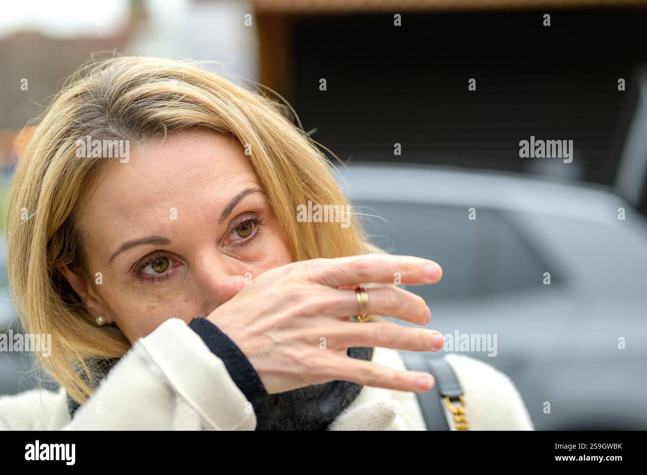 Eine Nahaufnahme einer Frau in einem beigefarbenen Mantel, die ihr Gesicht abwischt, mit einem nachdenklichen Ausdruck und zarten Details in ihren Augen Stockfoto