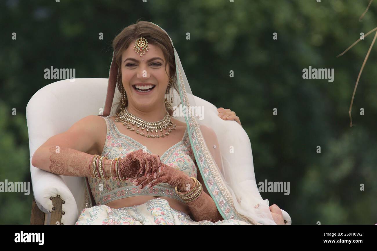 Schöne, fröhliche kaukasische Braut in indischer Brautkleidung lacht, während ihr Stuhl in der Tradition der hinduistischen Hochzeitszeremonie angehoben wird Stockfoto