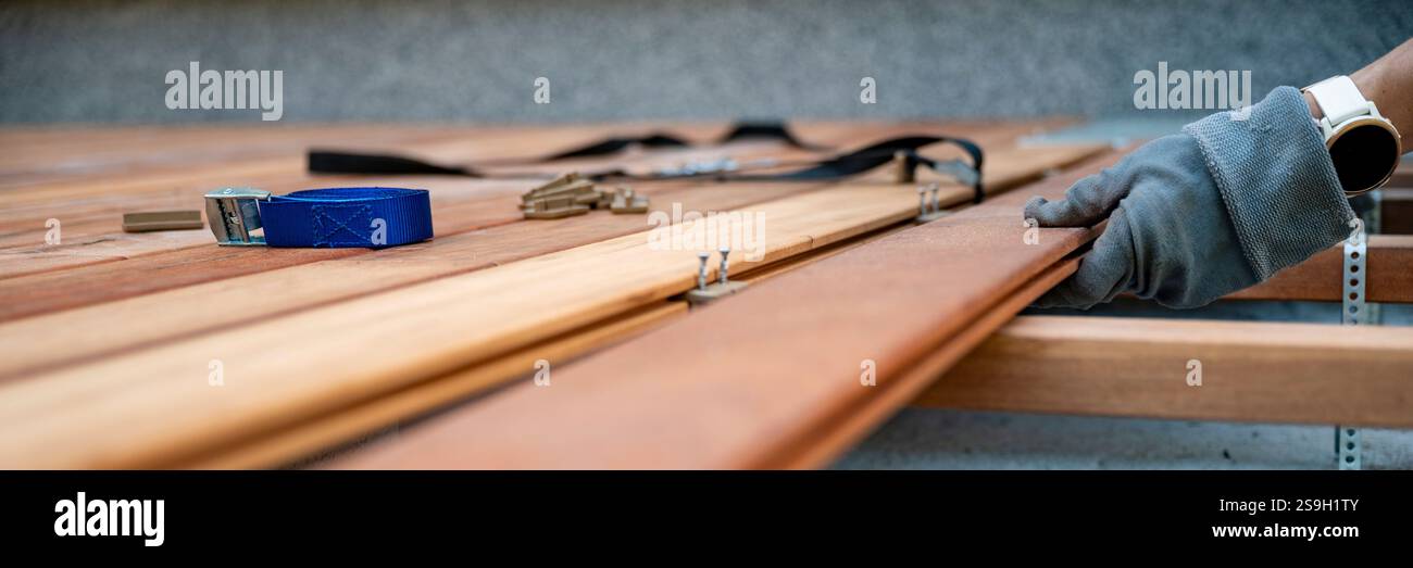 Eine Nahaufnahme eines Holzbearbeitungsprojekts, das eine Hand zeigt, die Holzdielen mit Handschuhen mit Werkzeugen wie einem blauen Ratschenriemen und Beschlägen ausrichtet, um die Preci hervorzuheben Stockfoto