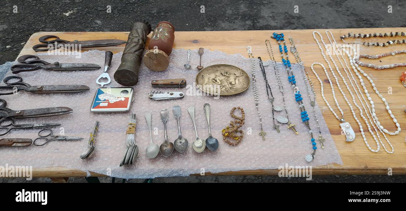 Europäischer Antiquitätenmarkt, Stand am Flohmarkt: Kristallschmuck, Bernsteinketten, massive barocke Perlenkette, Vintage Löffel, Silberlöffel, Vintage Rosenkranz Perlen, Naturstein und Mineral Halsketten, Edelstein Anhänger, Vintage Taschenmesser, Bronze Plakette, Vintage Gusseisen Scheren Sammlung, Graben Art Ein Gehäuse aus Messing aus dem Zweiten Weltkrieg Stockfoto