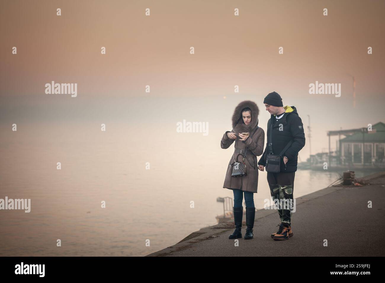 BELGRAD, SERBIEN - 12. JANUAR 2025: Ein Mann und eine Frau in Wintermänteln schlendern entlang der Donaupromenade, beobachten bei leichtem Nebel ein Smartphone, reflektieren Stockfoto