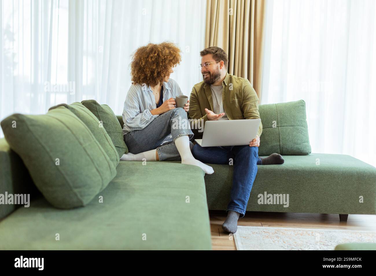 Ein fröhliches Paar sitzt nebeneinander auf einer weichen grünen Couch und führt ein aufregendes Gespräch, während es unter warmem Su auf einem Laptop Ideen durchstöbert Stockfoto