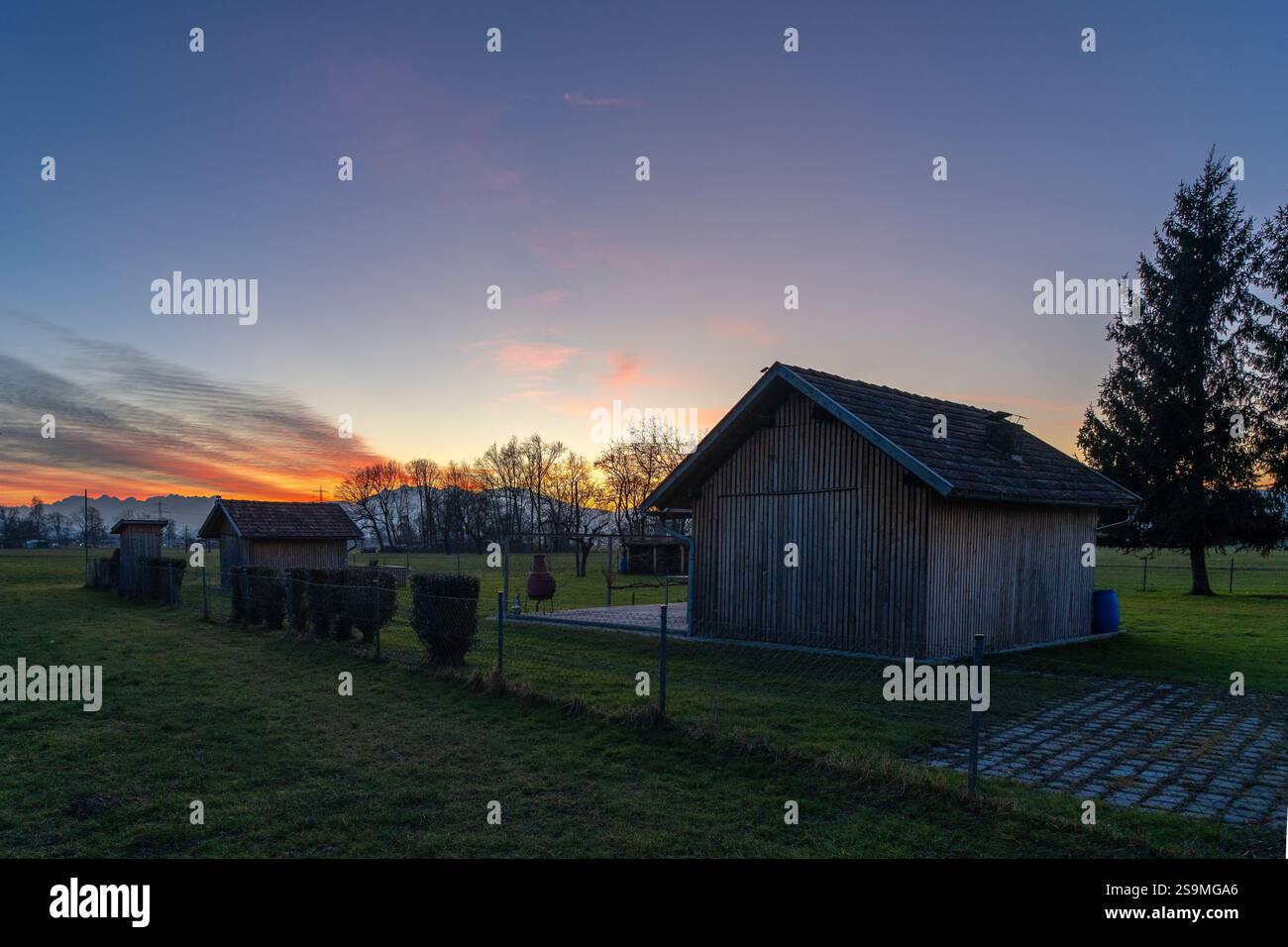 Nachglühpanorama mit Holzscheune auf Wiese, Garten und dominantem Baum unter Sonnenuntergang farbigem Himmel, mit orange, roten Wolken und blauem Himmel, Weide Stockfoto