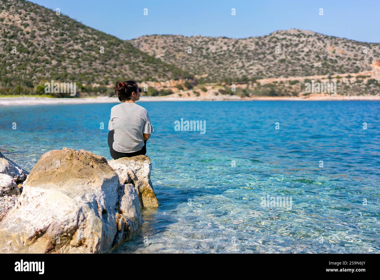 Rückansicht einer Frau, die auf einem Stein sitzt und auf einen Kiesstrand blickt Stockfoto