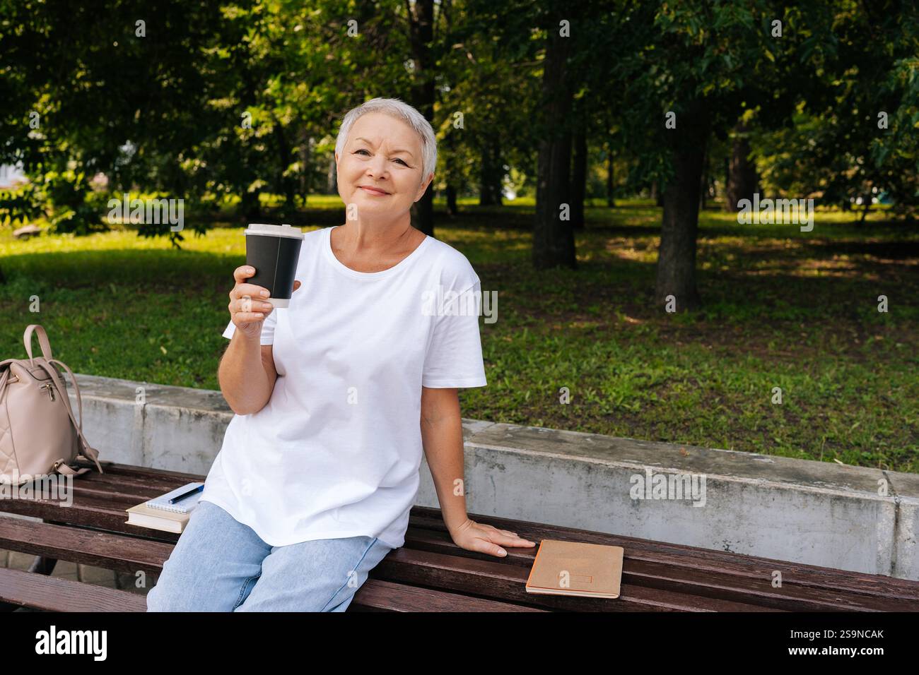 Porträt einer entspannten europäischen grauhaarigen älteren Frau, die auf der Bank sitzt, aus dem Becher schlürft und am Sommertag im öffentlichen Stadtpark im Freien genießt Stockfoto