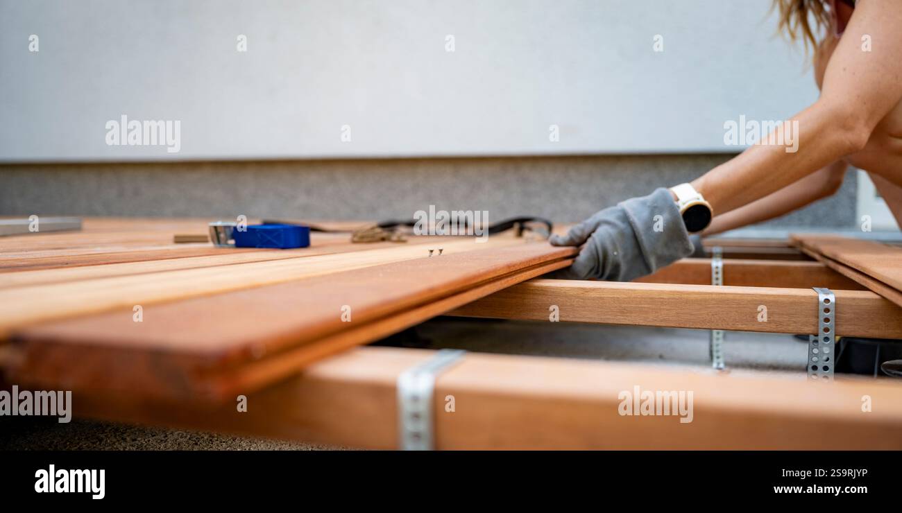 Frauenhände, Handschuhe und eine Smartwatch tragen, sorgfältig Holzdielen auf einem Deckrahmen während eines DIY-Projekts ausrichten. Stockfoto