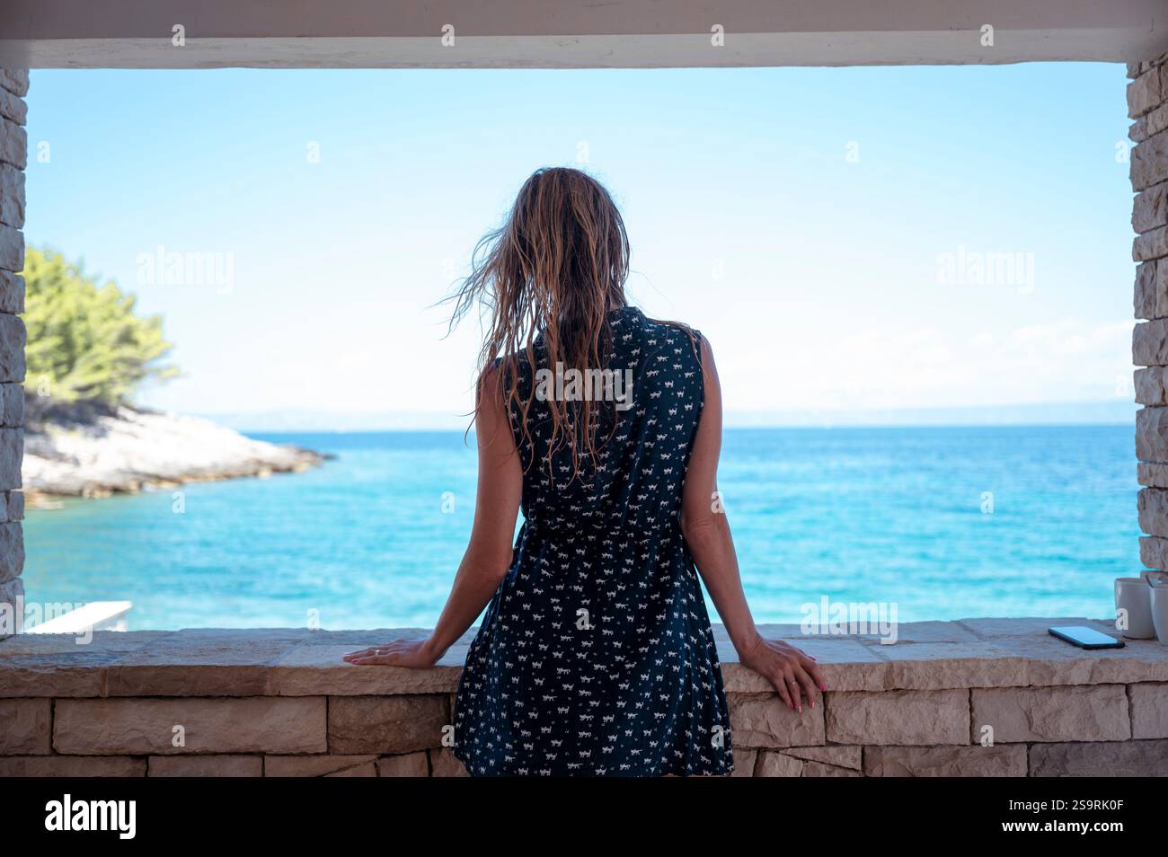 Eine Frau lehnt sich auf einem Steinbalkon und blickt auf das kristallklare blaue Meer und den Himmel, umgeben von einer ruhigen Küstenlandschaft und sanften Meeresbrisen in Summe Stockfoto