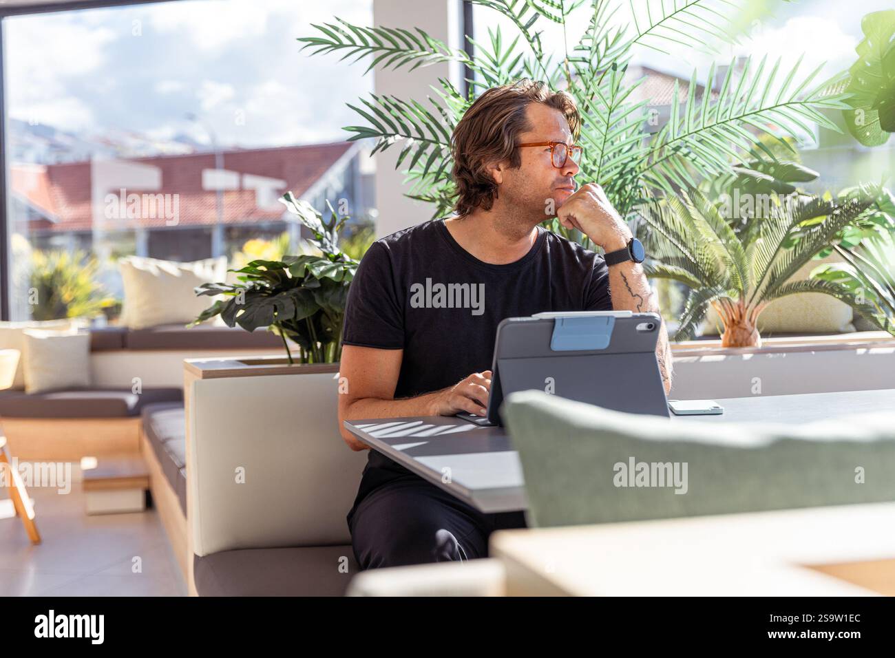 Ein Hipster-Geschäftsmann sitzt mit einem Tablet an einem Tisch und hat einen nachdenklichen Ausdruck. Stockfoto