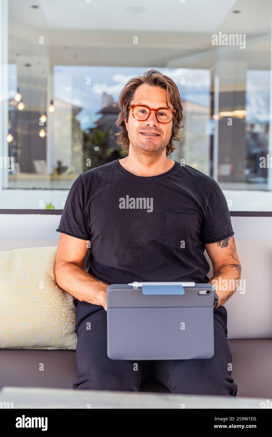 Das vertikale Porträt eines Mannes in einem schwarzen Hemd sitzt auf einer Couch und hält eine Tablette. Stockfoto