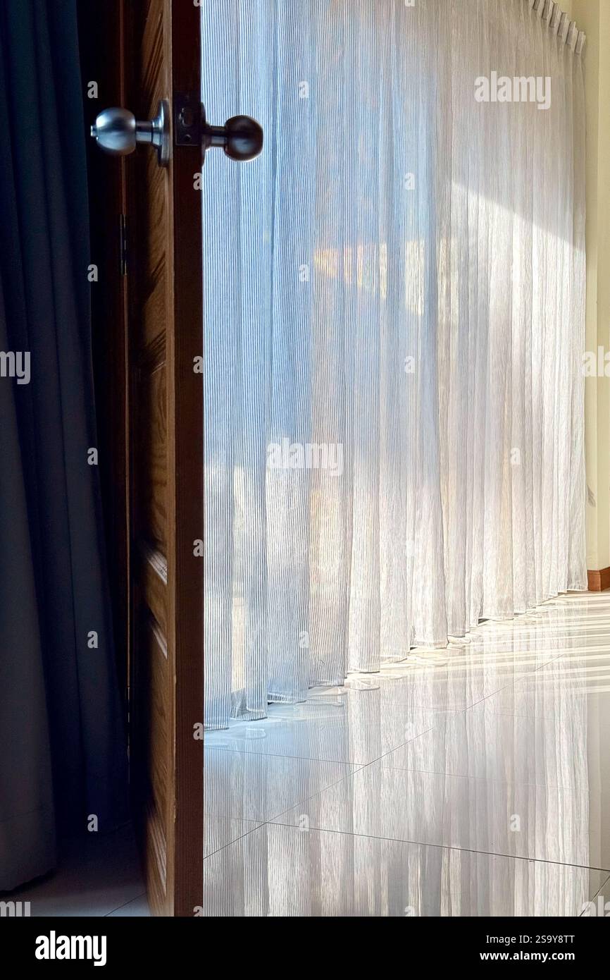 Einladendes Licht: Ein ruhiger Blick durch eine offene Tür Eine friedliche Szene Einer offenen Holztür, die zu einem sanft beleuchteten Raum mit transparenten Vorhängen führt. - Smartphone-aufgenommenes Stockfoto