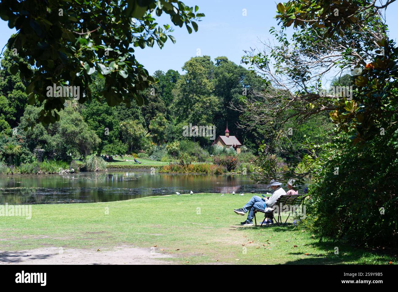 Ein asiatischer Mann und eine asiatische Frau sitzen und entspannen auf einem Platz am See unter blauem Himmel mit Blick auf das William Tell-Ruheraum, Royal Botanic Gardens Melbourne. Stockfoto