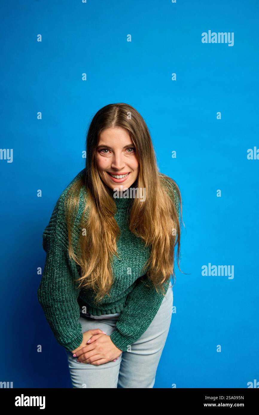 Studio-Aufnahme einer fröhlichen jungen Frau mit langen Haaren, die sich nach vorne beugt und auf einem leuchtend blauen Hintergrund lächelt Stockfoto