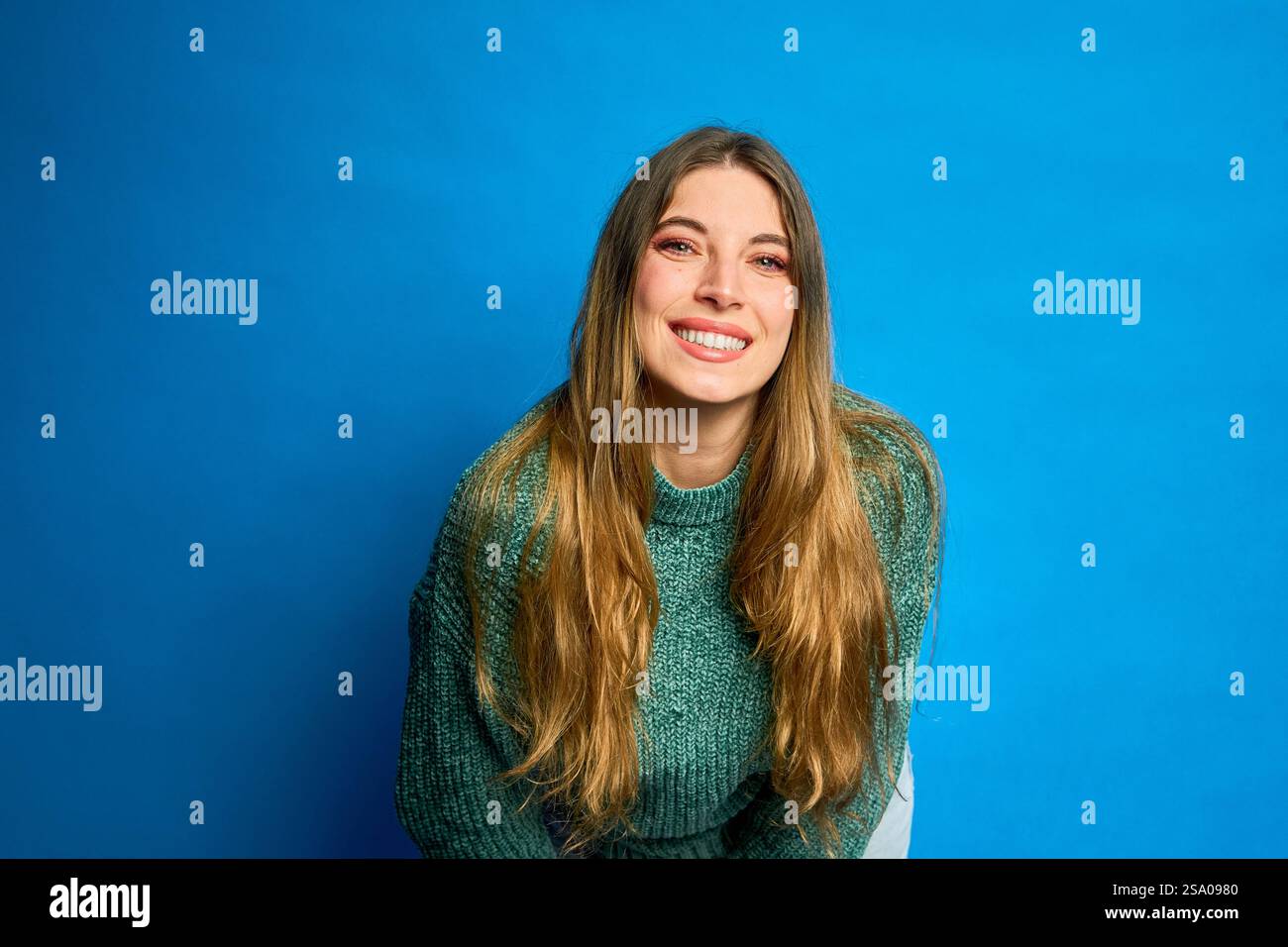 Studio-Porträt einer fröhlichen jungen Frau mit langen blonden Haaren, die grünen Pullover trägt, sich nach vorne lehnt und auf blauem Hintergrund lächelt Stockfoto