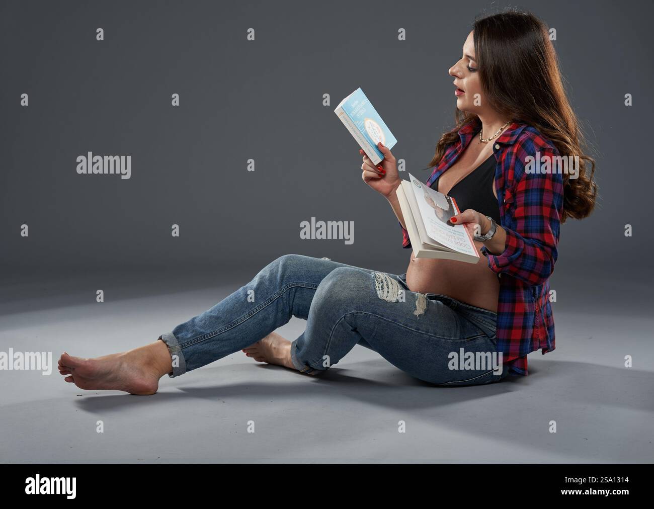 Eine schwangere Frau sitzt barfuß auf dem Boden und lächelt, während sie ein Buch über die Erziehung liest Stockfoto