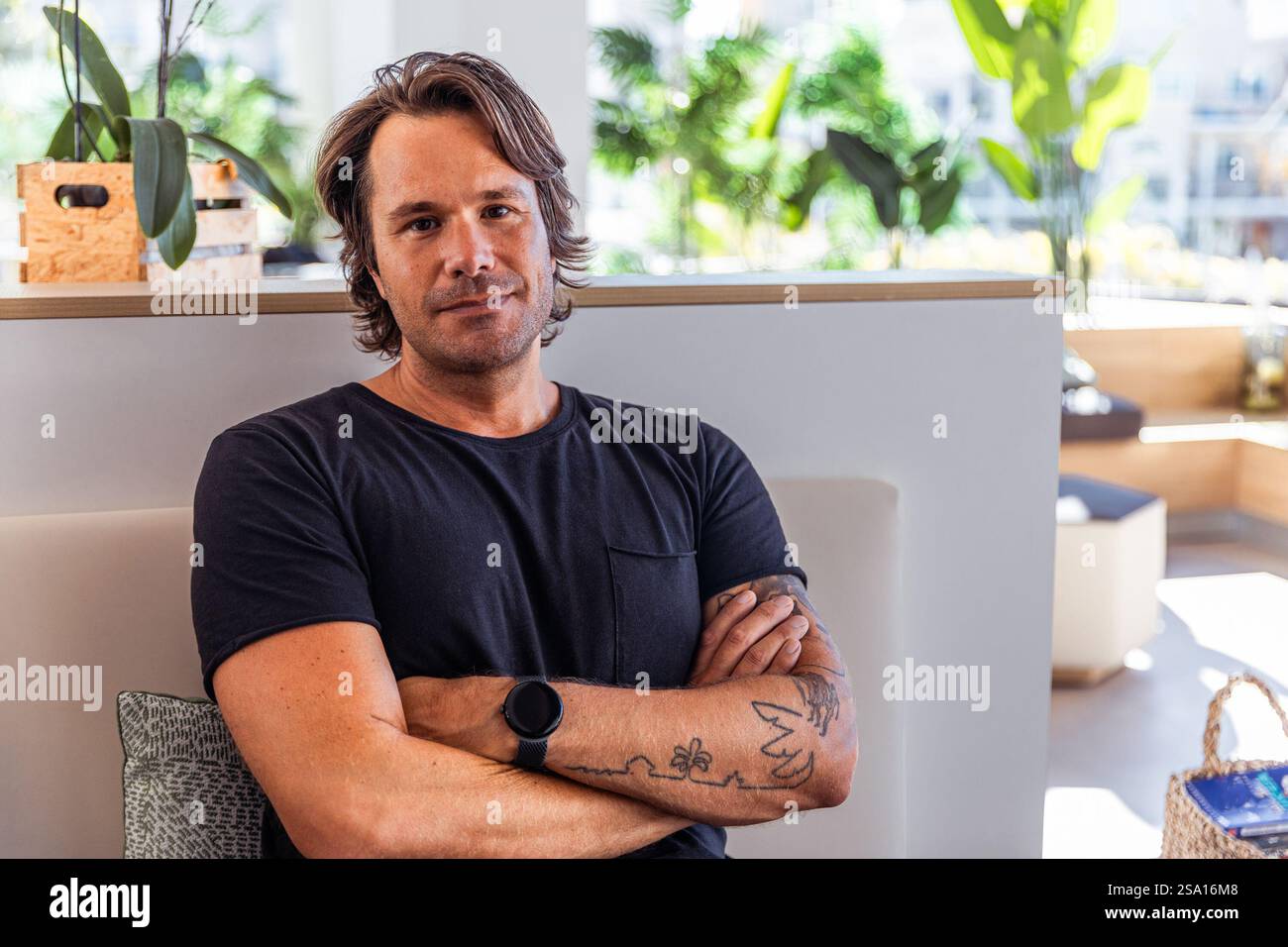 Ein selbstbewusster Mann mit einem schwarzen Hemd und einer Uhr sitzt mit überkreuzten Armen. Er hat ein Tattoo am Arm. Stockfoto