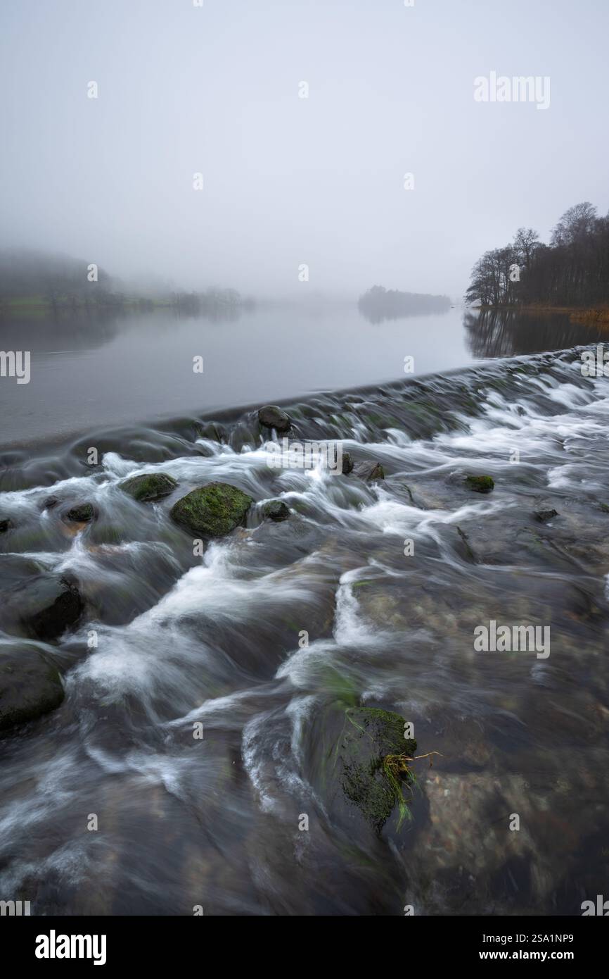 Fließende Wehr mit Blick auf einen nebeligen Grasmere See an einem nassen Wintermorgen im Lake District, Großbritannien. Stockfoto