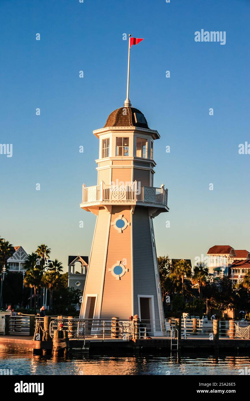 Ein Leuchtturm steht hoch auf einem Pier. Der Leuchtturm ist weiß und hat eine rote Flagge oben. Der Himmel ist blau und die Sonne untergeht Stockfoto