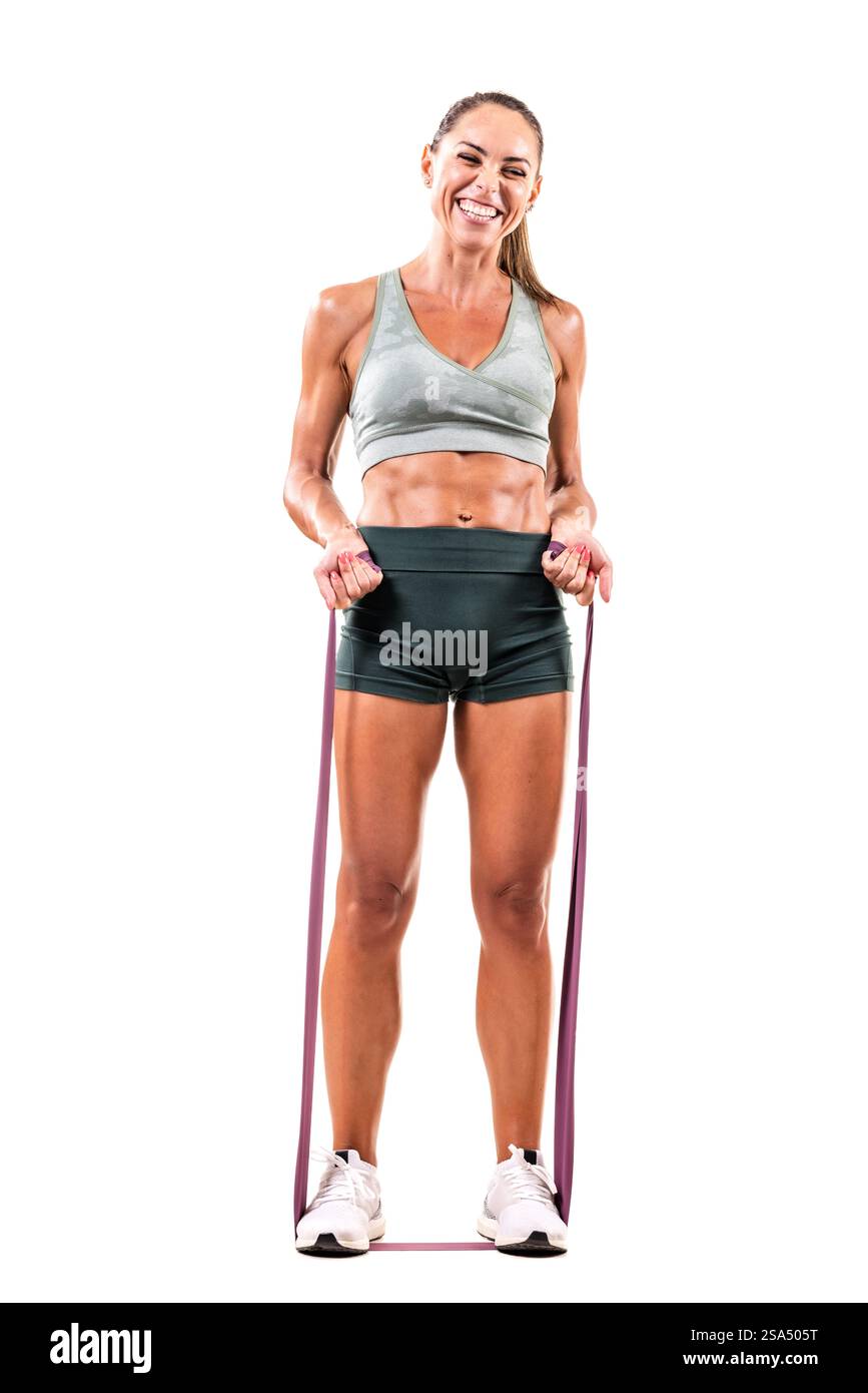 Eine Frau in sportlicher Kleidung steht lächelnd, während sie mit Widerstandsbändern vor weißem Hintergrund trainiert und zeigt Fitness, Kraft und Positivit Stockfoto