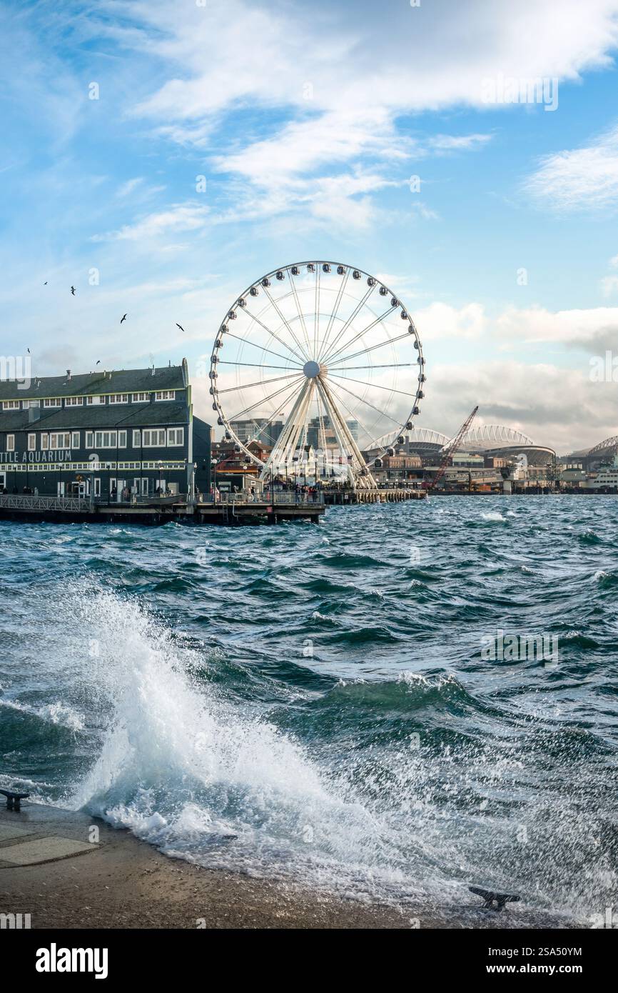 Das Seattle Great Wheel erhebt sich im Hintergrund, während die Wellen gegen das Ufer krachen und einen dynamischen und ikonischen Blick auf die Stadt schaffen. Stockfoto