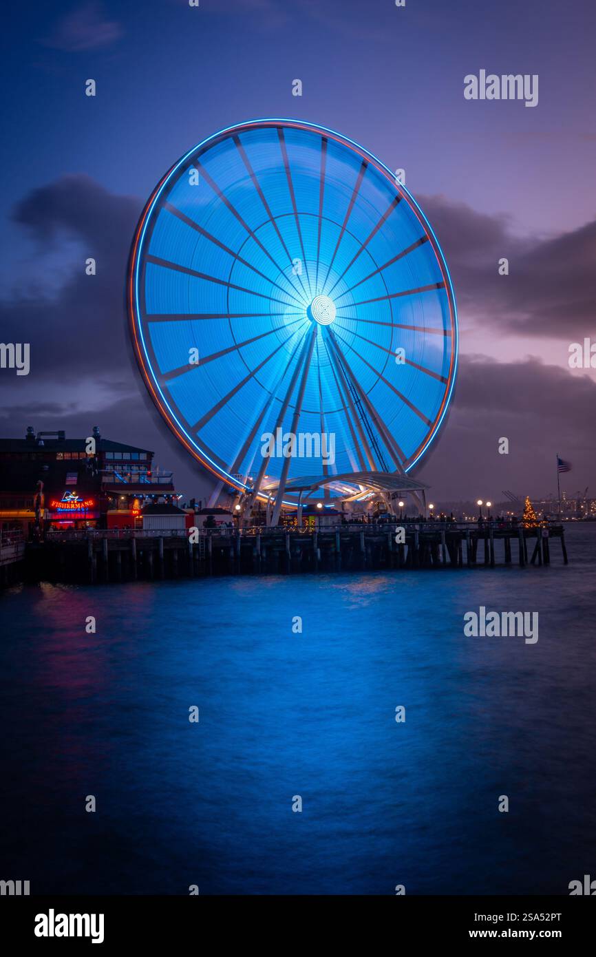 Ein lebhafter, bewölkter Sonnenuntergang zeigt das große Rad von Seattle, das am Abendhimmel und am Wasser leuchtet. Stockfoto