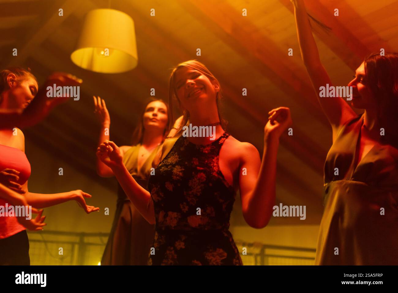 Junge Frauen tanzen fröhlich in einem schwach beleuchteten Raum und feiern Freundschaft und Freiheit. Die warme Atmosphäre trägt zum Gefühl von Glück und Jugendlichkeit bei Stockfoto