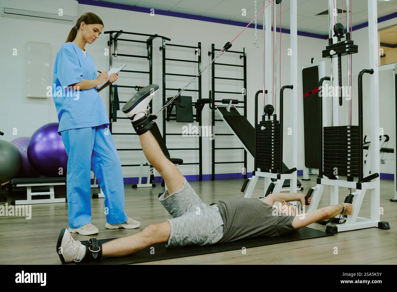 Tiefwinkelaufnahme eines jungen erwachsenen Physiotherapeuten zur Einschätzung der Fähigkeiten älterer Patienten, die auf der Matte liegen und Übungen für die unteren Gliedmaßen machen Stockfoto