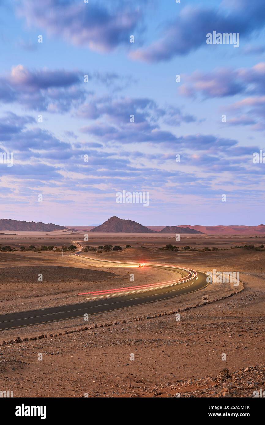 Leichte Pfade vorbeifahrender Autos auf der Straße nach Sossusvlei, frühmorgens blaue Stunde. Himmel mit flauschigen Wolken. Sesriem, Nationalpark, Namibia, Afrika. Stockfoto