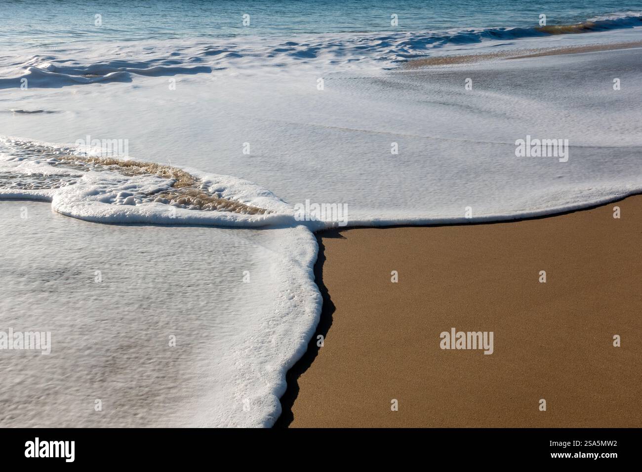 Sanfte Wellen gleiten über goldenen Sand und bilden komplizierte Designs, während die Sonne untergeht und ein warmes Leuchten über den ruhigen Strand wirft Stockfoto