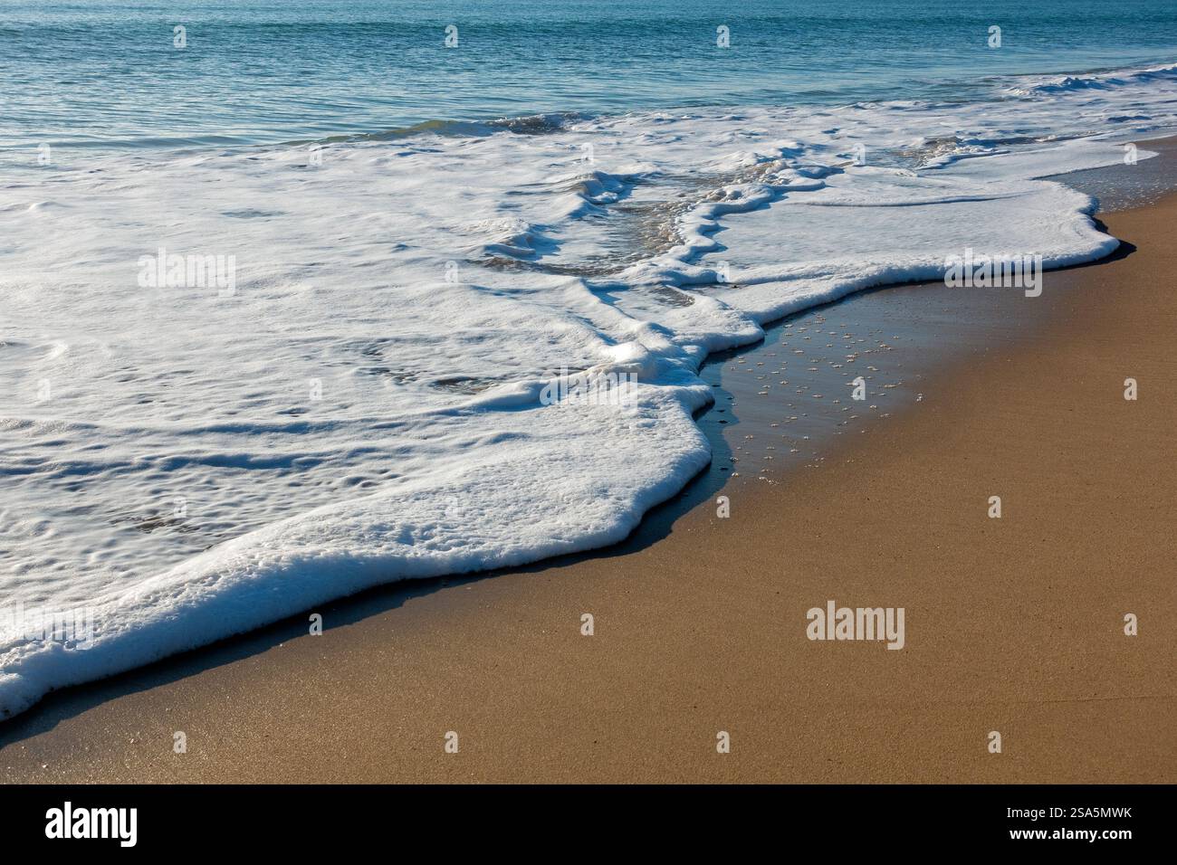 Sanfte Wellen gleiten über goldenen Sand und bilden komplizierte Designs, während die Sonne untergeht und ein warmes Leuchten über den ruhigen Strand wirft Stockfoto