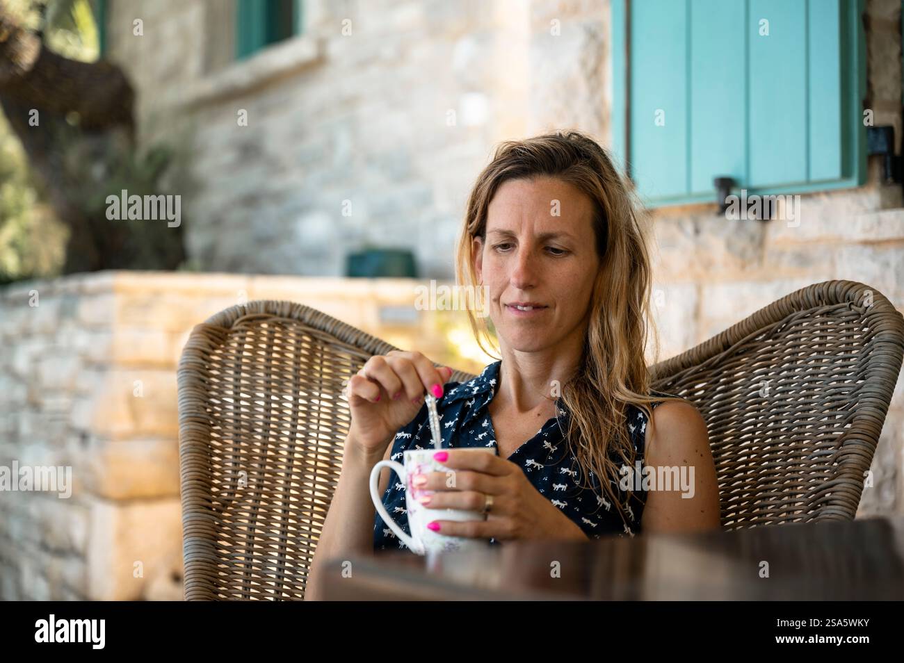 Junge Frau mit nassen Haaren, die in einem bequemen Stuhl mit einer Tasse Kaffee draußen auf einer Terrasse mit einem Steinhaus im Hintergrund sitzt. Freizeit va Stockfoto