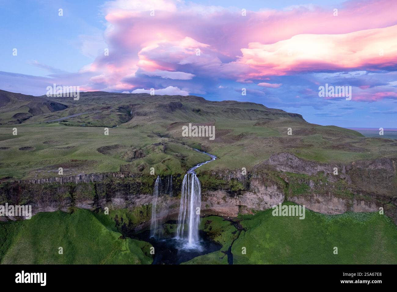 Aus der Vogelperspektive des majestätischen Seljalandsfoss Wasserfalls, der durch grüne Klippen unter leuchtenden Wolken bei Sonnenuntergang im Bereich des Golden Circle in Island kaskadiert Stockfoto