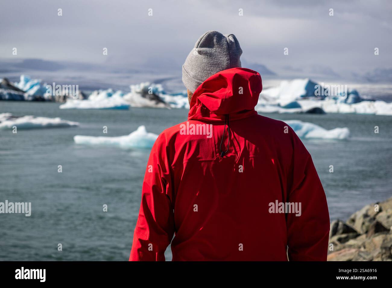 Reisende in roter Jacke, die die Jokulsarlon Lagune in der arktischen Landschaft Islands mit Eisbergen und Wolken beobachtet Stockfoto