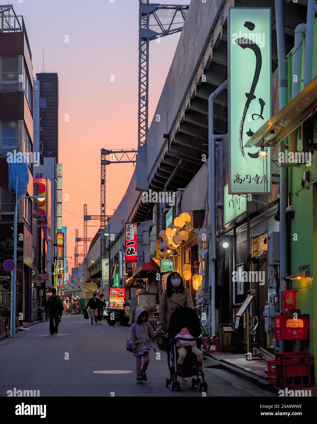 Blick am frühen Abend auf eine Straße, die neben den erhöhten Bahngleisen der Chuo-Linie im Westen Tokios verläuft, mit mehreren Fußgängern und Geschäften Stockfoto