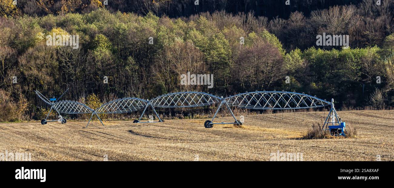 Ein großes Bewässerungssystem erstreckt sich über ein ausgeerntetes Feld, das vor dem Hintergrund von herbstfarbenen Bäumen positioniert ist. Die Ausrüstung ist bereit für die Zukunft Stockfoto