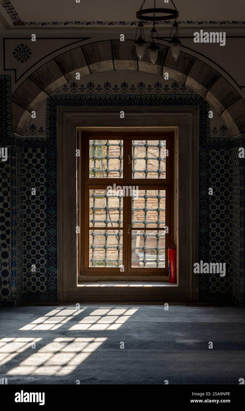 Reich verzierte Fenster mit komplizierten Holzgittern und traditionellen osmanischen Fliesen in einer historischen Moschee. Sonnenlicht wirft geometrische Schatten auf den Boden Stockfoto