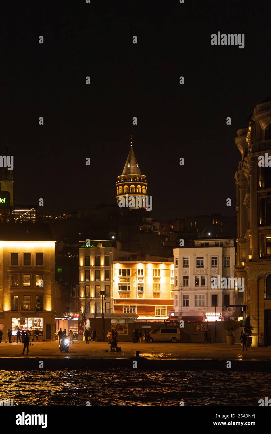 Nachtblick auf den beleuchteten Galatenturm und einen modernen Uhrenturm in Istanbul. Die warmen Lichter stehen im Kontrast zum dunklen Himmel Stockfoto