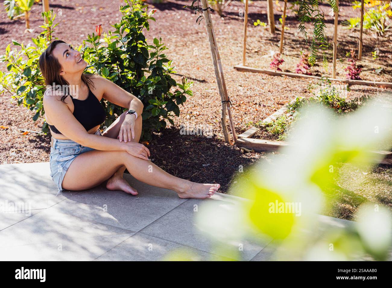 Eine Frau sitzt auf dem Boden in einem Garten. Sie trägt ein schwarzes Tanktop und Shorts Stockfoto