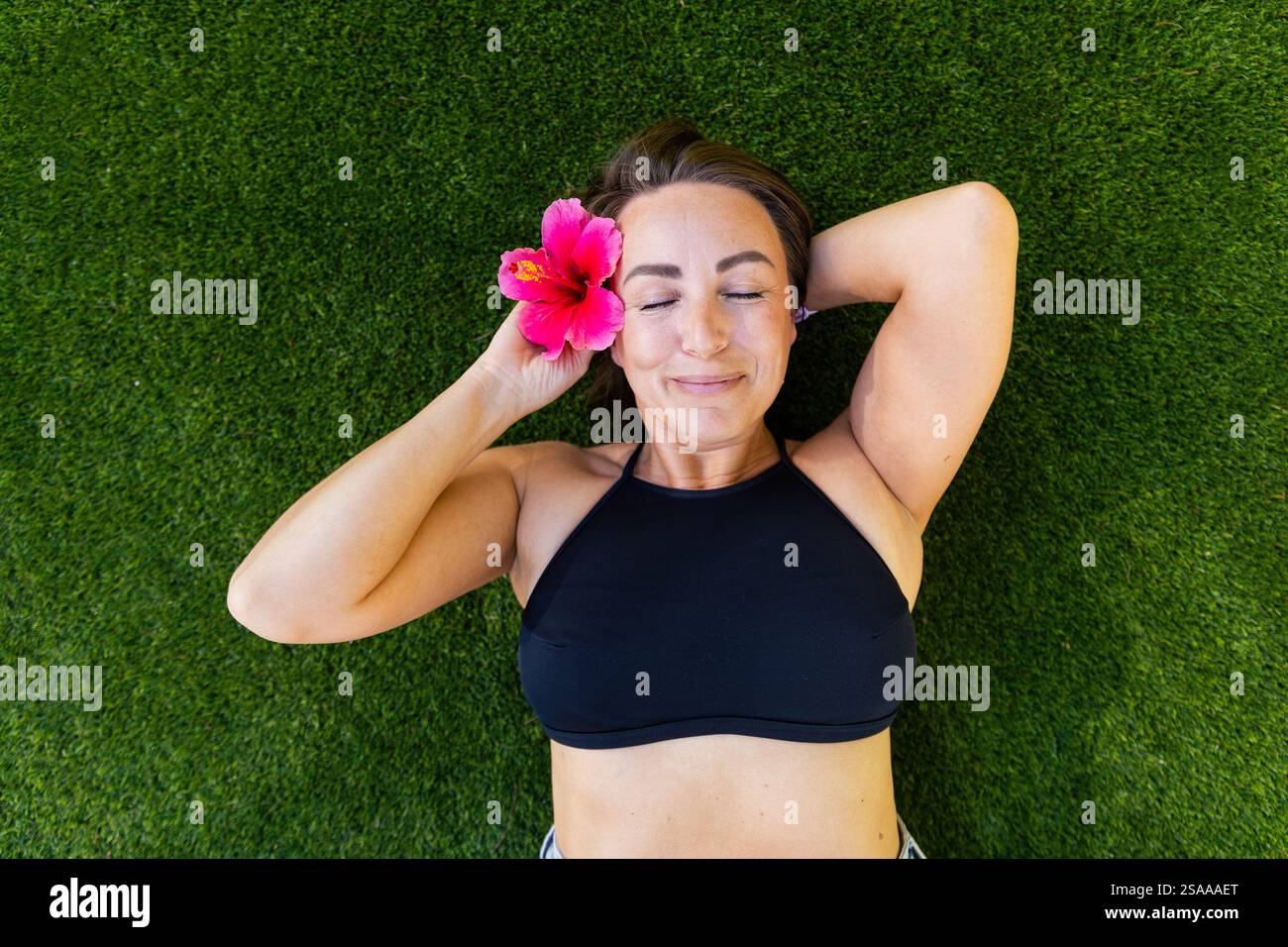 Eine Frau liegt auf dem Gras mit einer rosa Blume im Haar und geschlossenen Augen. Sie lächelt und ist entspannt Stockfoto