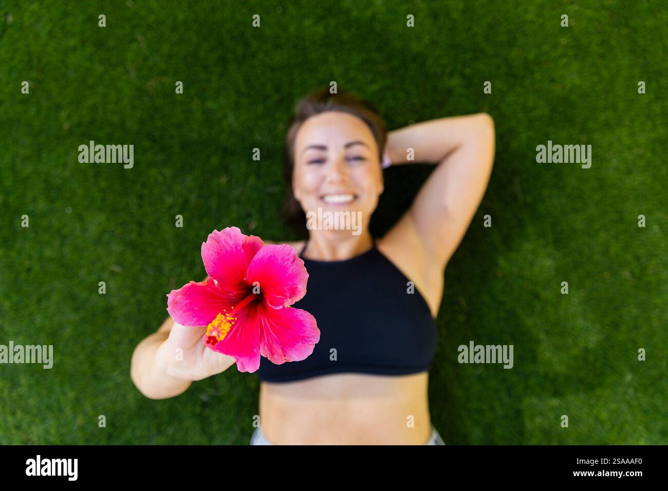 Eine Frau liegt auf dem Gras und hält eine rosa Blume. Sie lächelt und genießt den Moment Stockfoto